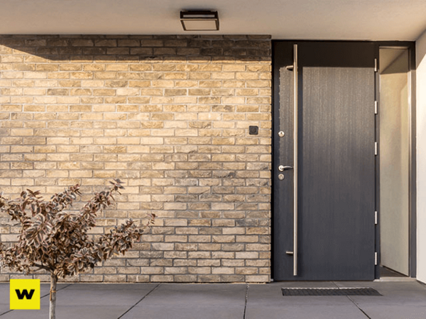 Moderne Haustür aus dunklem Material in einer hellen Klinkerfassade mit seitlicher Pflanze.